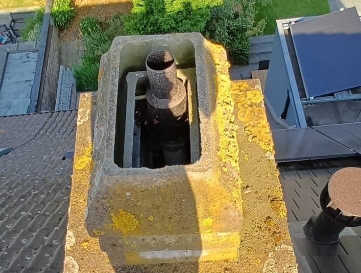 Concrete chimney seen from above, covered in yellow lichen, among dark tiles, in Grimbergen