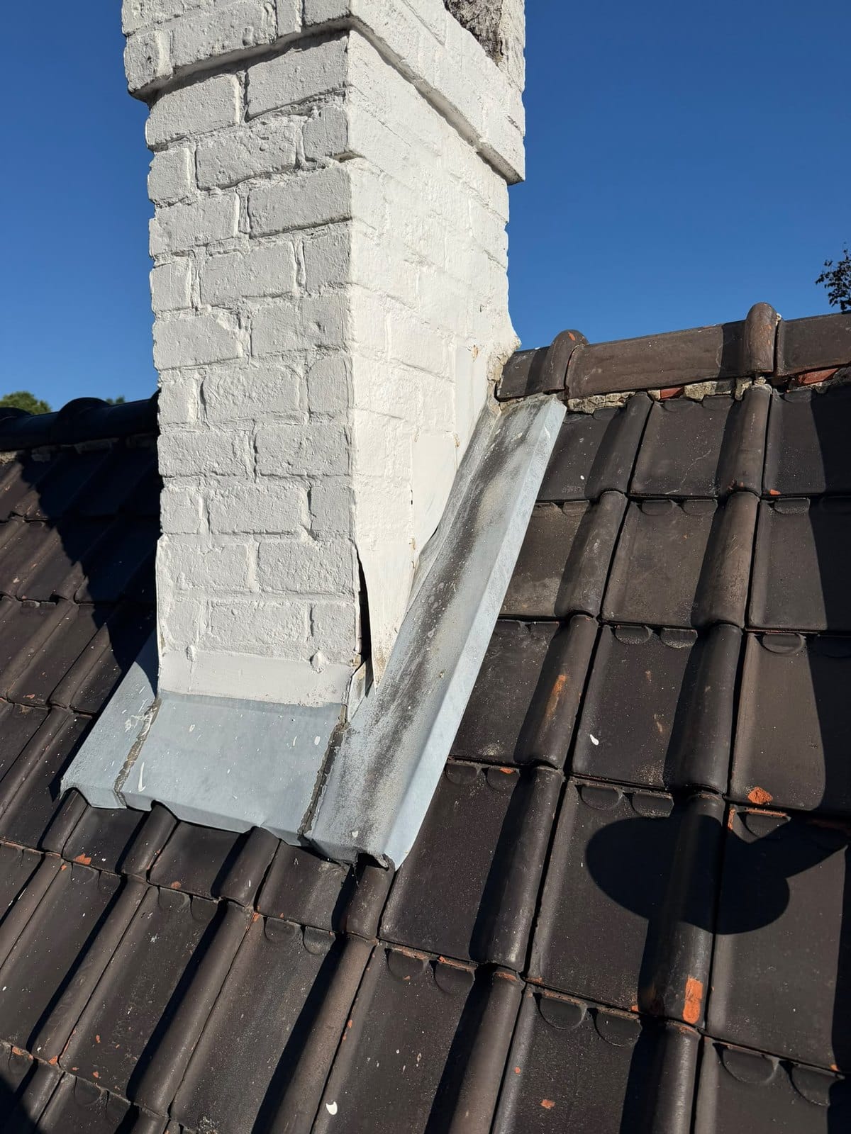Brick chimney painted white with a zinc strip along the slope, surrounded by brown tiles, under a blue sky