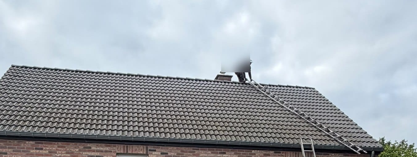 Two-slope roof of grey tiles under an overcast sky, roof ladder, a team member with a blurred face near the chimney