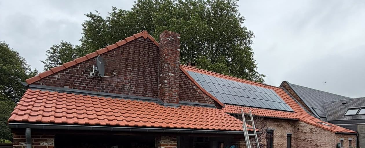 Toiture en tuiles orange vif, cheminée en briques et panneaux solaires sur le versant voisin, échelle appuyée contre la gouttière, ciel couvert
